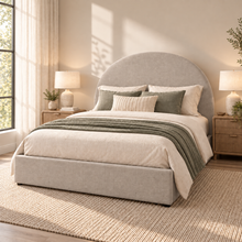 Load image into Gallery viewer, A modern upholstered bed with a tall rounded headboard and matching base, styled in a bright neutral bedroom. The bed is dressed in cream bedding, green accent pillows, and a textured green throw blanket, flanked by two wooden nightstands with lit table lamps.