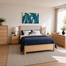 Load image into Gallery viewer, A modern bedroom featuring a light oak wood bed frame with a beige upholstered headboard, styled with a navy blue quilt and white pillows. The room includes matching light oak nightstands, a tall dresser, and a matching chest of drawers, all set on a large woven area rug.