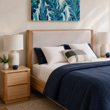 Load image into Gallery viewer, A styled bedroom scene showing a bed with a light wood frame and a beige upholstered headboard, dressed in white and navy bedding with decorative pillows. Matching nightstands with white lamps sit on both sides, and a colorful tropical leaf wall art piece hangs above the bed.