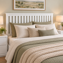 Load image into Gallery viewer, A neatly made bed in a softly lit bedroom, featuring a white slatted headboard, layered cream and sage green pillows, a cream knit blanket, and a chunky sage green throw. Nightstands with lit lamps and a framed landscape print are visible.
