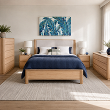 Load image into Gallery viewer, A styled bedroom scene featuring a light oak wood bed frame with a beige upholstered headboard, dressed in a navy blue quilt and white pillows. The bed is flanked by matching light oak nightstands with white table lamps, and the room is completed with a matching dresser, a textured beige rug, and a blue and white tropical leaf wall art print.