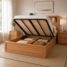 Load image into Gallery viewer, A light oak wood platform storage bed with its lift-up headboard panel raised, showing the black metal support frame and storage compartments underneath. The bed is styled in a warm, neutral-toned bedroom with matching nightstands, a lamp, and a textured rug.