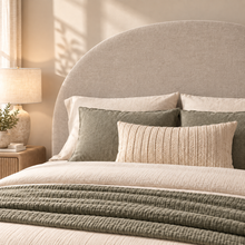 Load image into Gallery viewer, A styled bed in a softly lit bedroom featuring a large upholstered headboard, cream bedding, sage green pillows, and a matching textured throw blanket. A bedside lamp and small plant are visible on a wooden nightstand.