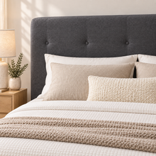 Load image into Gallery viewer, A dark gray upholstered tufted bed headboard with white and beige bedding, a textured cream pillow, and a beige knit throw blanket, styled in a warm, modern bedroom setting next to a wooden nightstand with a lamp.
