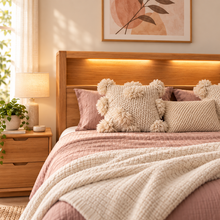 Load image into Gallery viewer, A cozy bedroom scene with a wooden bed frame, blush pink bedding, textured cream throw pillows, and warm ambient lighting from a bedside lamp and wall sconce.