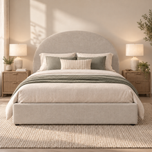 Load image into Gallery viewer, A neatly made bed with a tall upholstered headboard and matching footboard, styled in a warm neutral bedroom with two bedside tables, lamps, plants, and a textured rug.