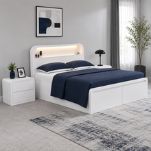Load image into Gallery viewer, A modern white wooden bed frame with an integrated backlit headboard, dressed in navy blue bedding, shown in a styled bedroom setting with a matching nightstand, rug, and window.