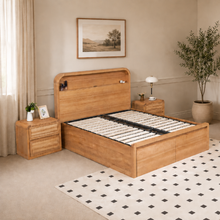 Load image into Gallery viewer, A light oak wood bed frame with a matching headboard and footboard, shown in a styled bedroom setting with a matching nightstand, lamp, rug, and potted plant.