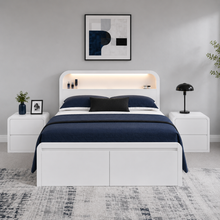 Load image into Gallery viewer, A modern white platform bed with a matching headboard and footboard, styled in a bedroom setting with navy blue bedding, white nightstands, and a gray rug.