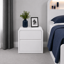 Load image into Gallery viewer, A modern white two-drawer nightstand in a bedroom setting, styled with a dark blue vase containing green leaves and a black-framed picture frame, next to a bed with navy bedding.