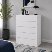 Load image into Gallery viewer, A white five-drawer chest of drawers in a modern bedroom setting, styled with a small potted plant, a black desk lamp, and a framed picture on top.