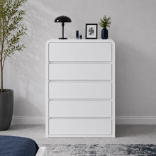 Load image into Gallery viewer, A white five-drawer chest of drawers stands against a light gray wall in a styled bedroom setting, with a black table lamp, small framed picture, and a dark blue vase on top. A potted plant and part of a bed are visible around it.