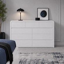 Load image into Gallery viewer, A modern white wooden dresser with six drawers, styled in a bedroom setting with a black table lamp, a framed print, and a blue bed visible in the foreground.