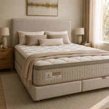 Load image into Gallery viewer, A fully made bed in a softly lit bedroom, featuring a quilted mattress with a beige upholstered base, matching pillows, a cream quilted cover, and a folded beige throw blanket. Nightstands, lamps, curtains, and wall art surround the bed.