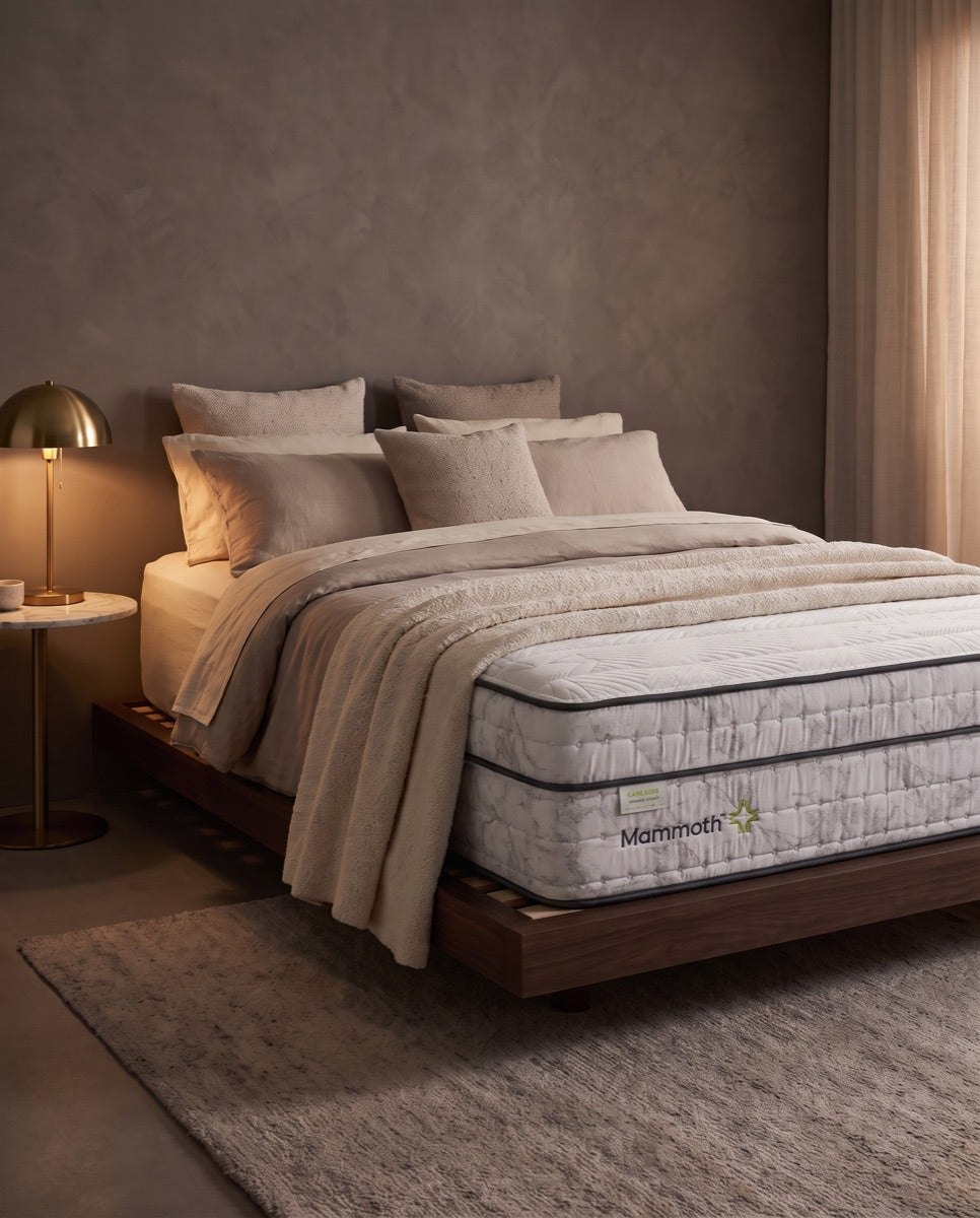 A bed with a white tufted headboard and a white mattress branded 'Mehndelian' is styled in a bright, modern bedroom with beige walls, a window, curtains, and a textured rug.